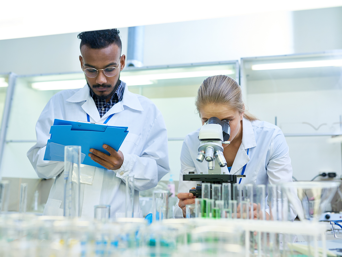 Man writing in a lab while woman looks through microscope