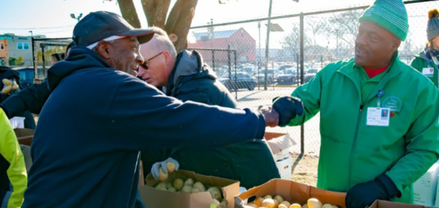 A male volunteer in a green hat and jacket shakes the hand of another man