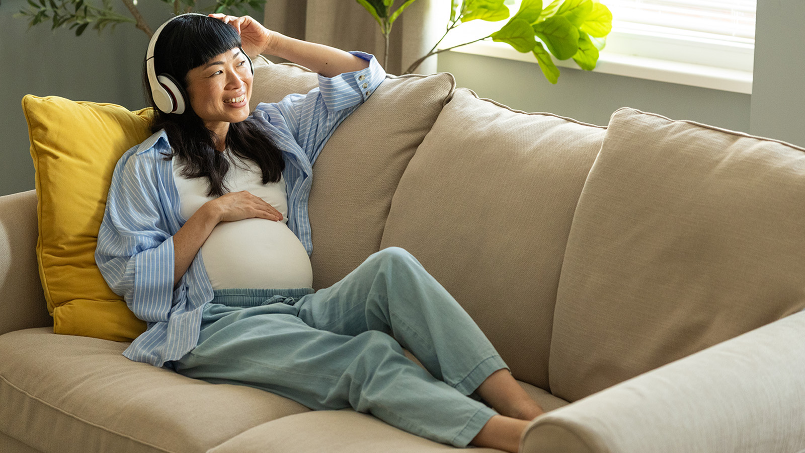 Pregnant woman relaxing on couch with her hand on her belly, smiling while looking out window and wearing headphones