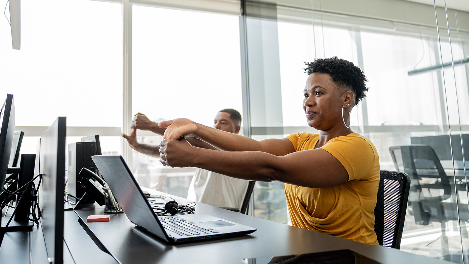Young woman and man doing wrist stretches at their desk in front of their computers in an office with large windows.