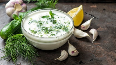 bowl of cucumber yogurt tzatziki dip with lemon wedge, cucumber, garlic, and mint springs on a wooden table