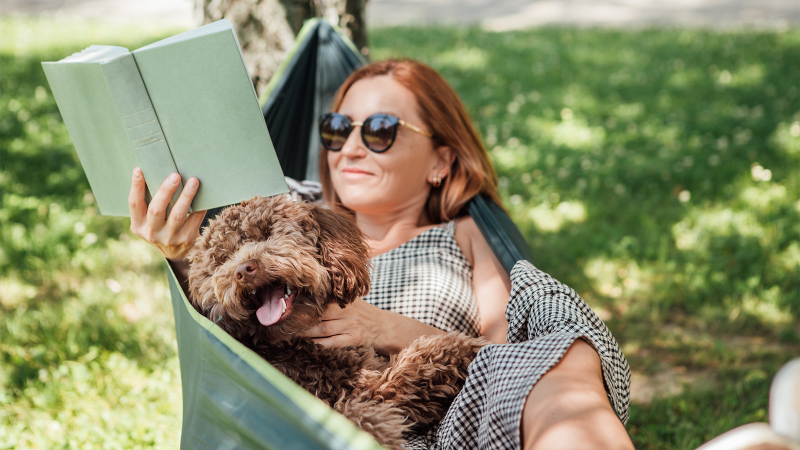 Woman wearing sunglasses and reading a book while relaxing in a hammock with a brown dog