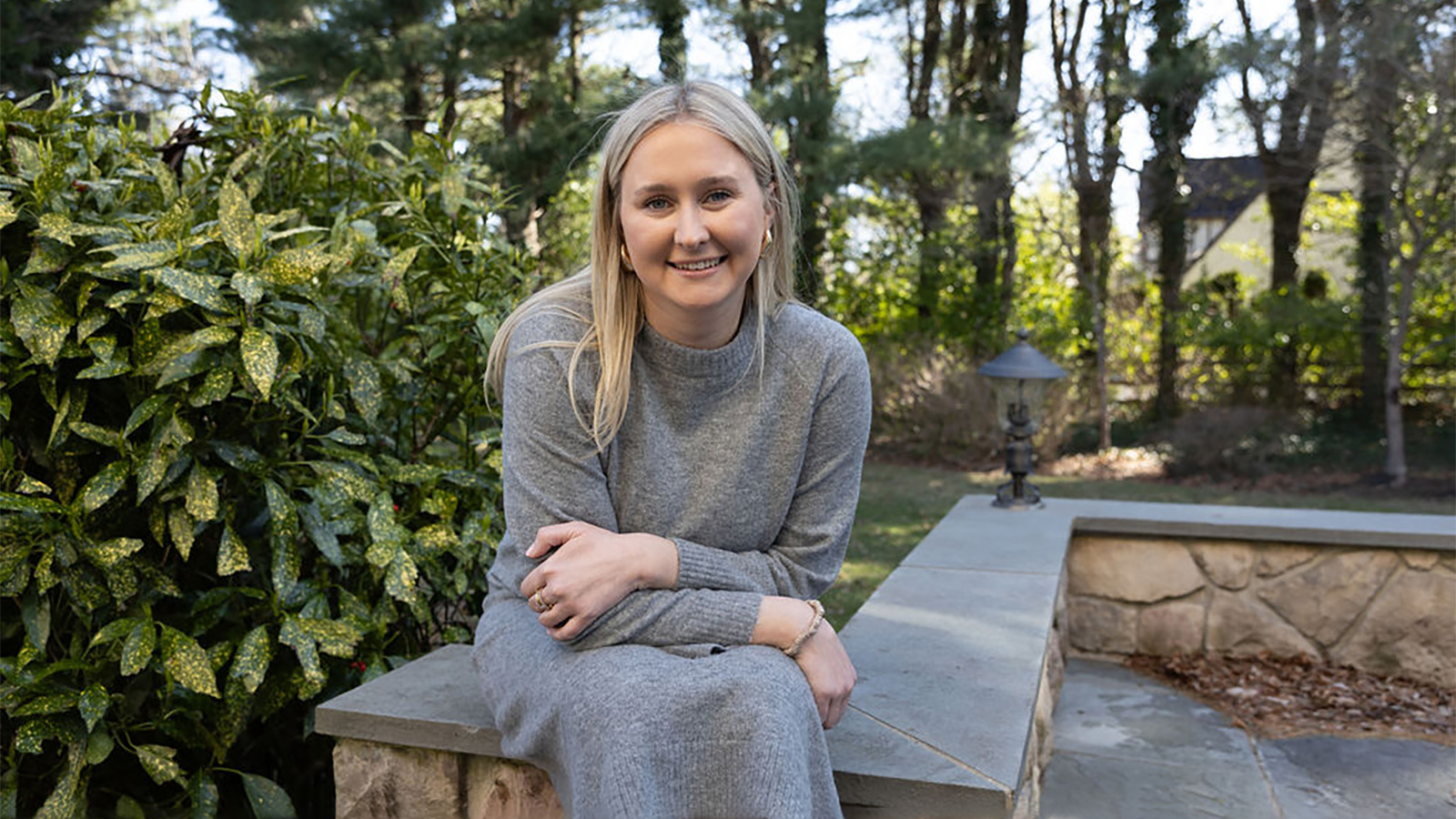 Woman sitting on a stone ledge in a park setting with trees behind her