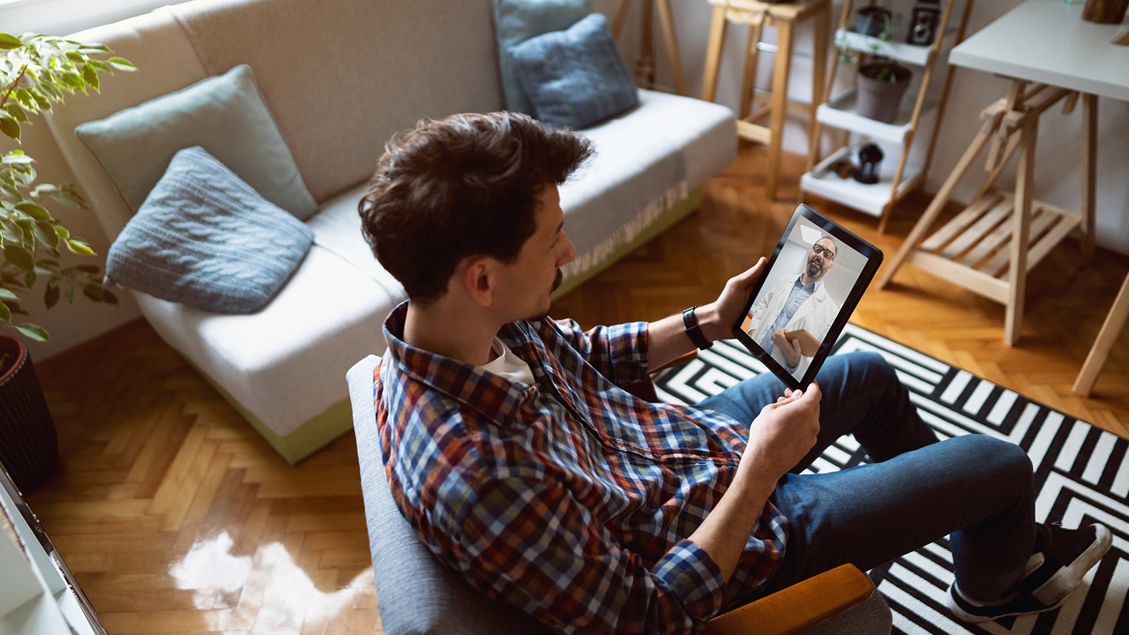 Young adult male sits on a chair and has a telehealth visit with a doctor on a tablet.