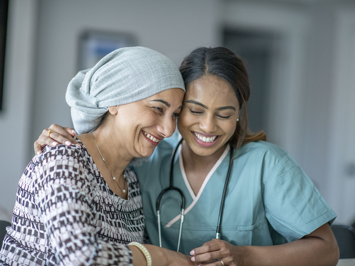 An older adult woman cancer patient embraces a female nurse in green scrubs.