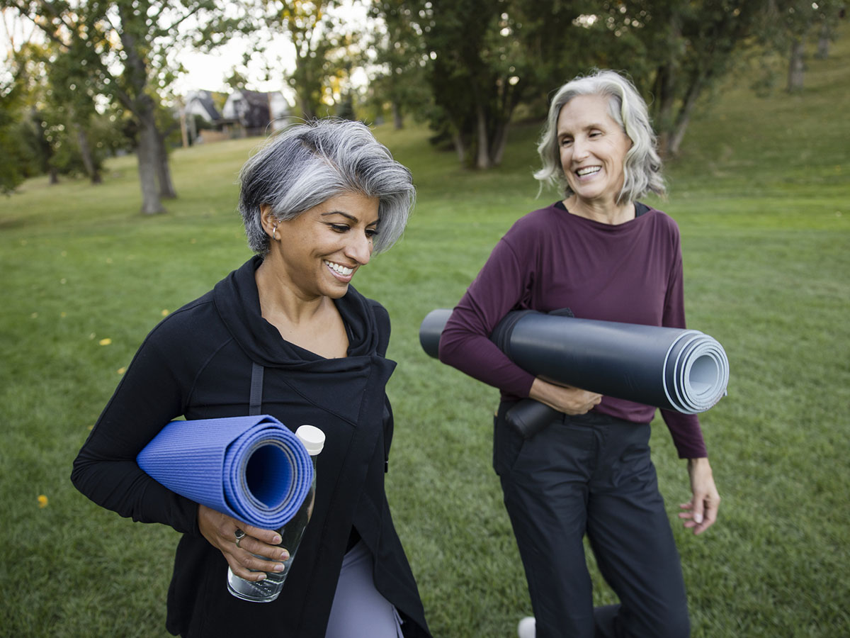 Two older adult women hold their yoga mats as they walk on the lawn.